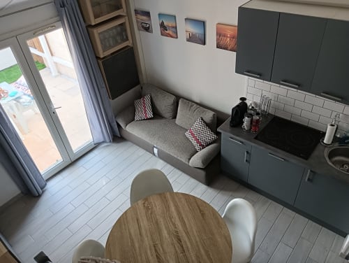 Salon du Gîte des Gônes avec canapé gris, table à manger, kitchenette, TV et vue sur le jardin par la terrasse