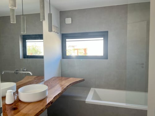 Banheiro moderno com bancada de madeira, cuba branca, paredes com azulejos cinza, box de vidro e janelas com vista