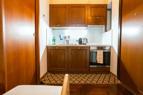 Cucina compatta in monolocale romano con mobili in legno, rivestimento bianco, forno integrato e tappetino decorativo.