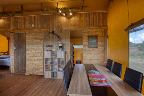 Modern glamping interieur met houten huisstructuur, eettafel, gele muren en uitzicht op het platteland bij Home Sweet Lodge