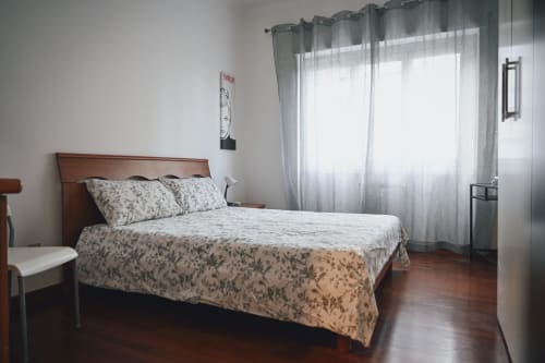 Camera da letto luminosa a Roma con letto in legno, biancheria floreale e ampie finestre con tende sheer