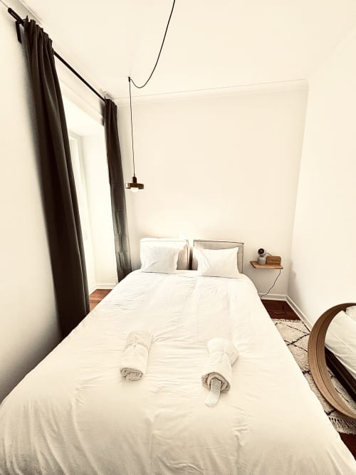 Minimalist bedroom in Royal Prince Garden Apartments, Lisboa, with white bedding, dark curtains, pendant light, and wooden