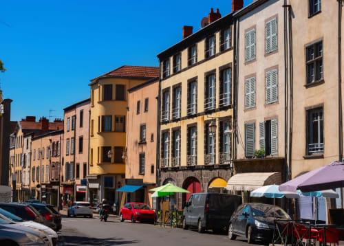 Colorful historic buildings lining a sunny square in Riom, France, with market stalls and parked cars below.