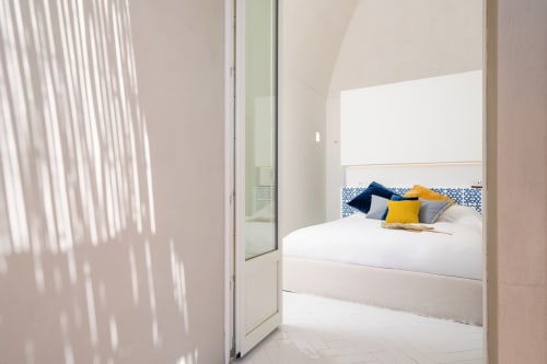 Camera da letto moderna e luminosa a La Conciergerie Capri con biancheria bianca, testata blu fantasia e cuscini colorati