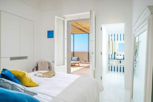 Camera luminosa a La Conciergerie Capri con vista sul mare attraverso porte in vetro, biancheria bianca, accenti blu e accesso alla terrazza.