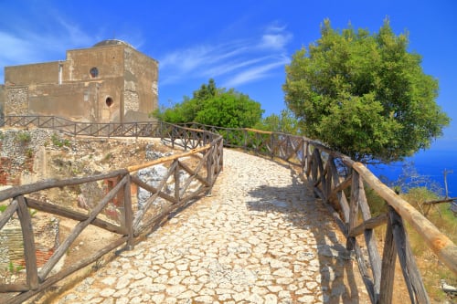 Antico sentiero in pietra con ringhiere in legno che conduce a una torre millenaria a Capri, circondata dalla verdeggiante macchia mediterranea e dal mare