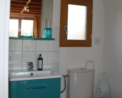 Modern bathroom in Domaine des Piéris cottage with turquoise vanity, white tiles, wooden window, and toilet