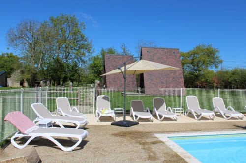 Poolside lounge area at Domaine des Piéris with white sunbeds, umbrella, and brick cottage in Grandcamp-Maisy