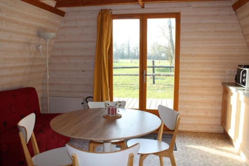 Bright wooden cottage dining area with round table, red sofa, and garden views through French doors at Domaine des Piéris