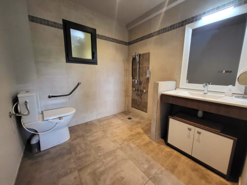 Spacious accessible bathroom at Villas Lantana with walk-in shower, white toilet, vanity, and dark tile flooring in Lecci.
