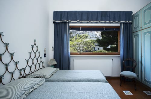Camera doppia a La Conciergerie Capri con tende blu scuro e finestra in legno affacciata sui giardini di Capri