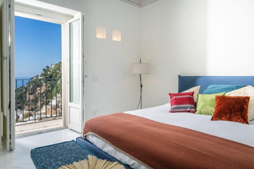 Camera luminosa con vista mare dal balcone, cuscini colorati e paesaggio della scogliera costiera a La Conciergerie Capri