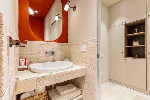 Moderno bagno presso Petra Apartments Roma con piastrelle in travertino beige, parete accent rossa, specchio rotondo e lavabo bianco