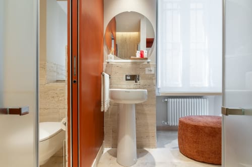 Bagno moderno alle Petra Apartments Roma con lavabo su piedistallo, specchio rotondo e rivestimenti in travertino