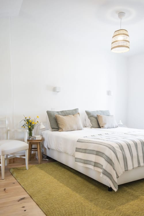 Quarto claro na Casa das Jardas com paredes brancas, roupa de cama listrada, luminária pendente tecida e tapete amarelo em Ladoeiro
