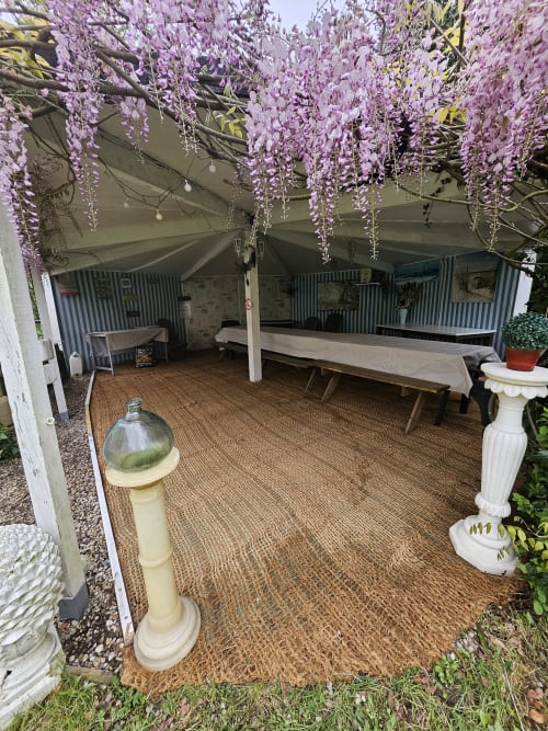 Überdachter Essbereich im Freien des Cottage Le Manoir Lacustre in Queyrac mit violetten Glyzinienblüten und Holzbänken