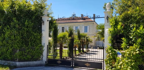 Eingang des Cottage Le Manoir Lacustre in Queyrac mit Toreinfahrt, Palmen und gepflegten Gärten unter blauem Himmel