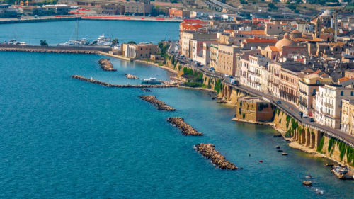 Coastal waterfront cityscape with turquoise Mediterranean waters, historic buildings, and moored yachts in Italy