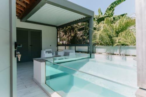 Piscine turquoise moderne avec parois vitrées, salon de détente, pergola verte et plantes tropicales à l'Habitation I'SEO