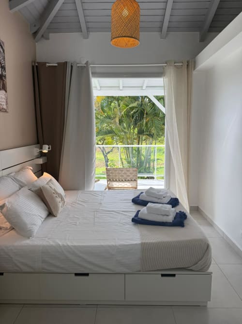 Chambre moderne et lumineuse au Domaine de la Glacière avec literie blanche, suspension en rotin et vue sur palmiers tropicaux
