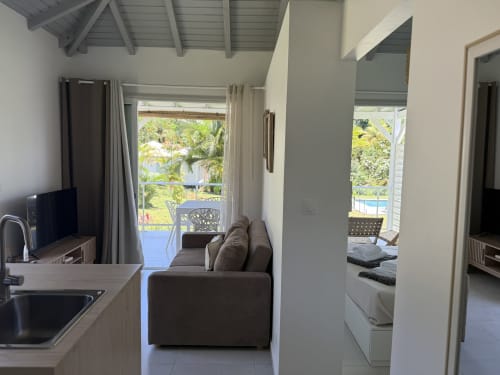 Séjour du bungalow Alpinia à la Domaine de la Glacière avec canapé gris, terrasse vue piscine et cuisine moderne à Petit Bourg,