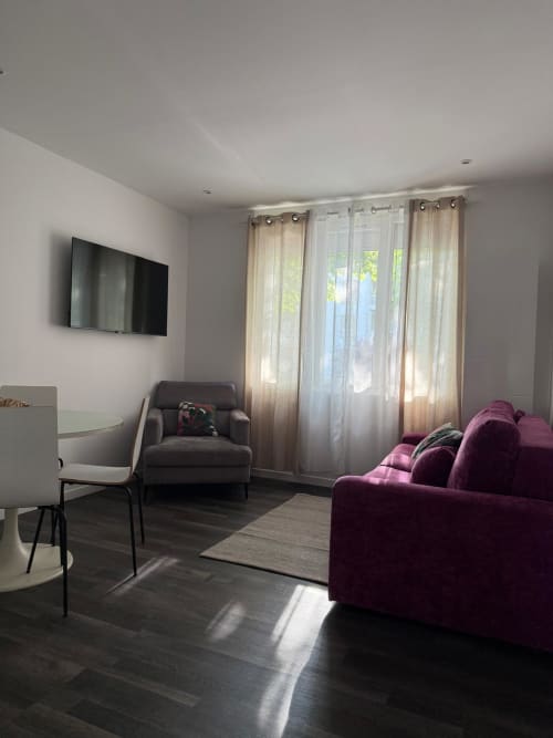 Élégant salon d'appartement hôtel Panambi avec canapé bordeaux, TV et vue sur la ville par des rideaux voilages