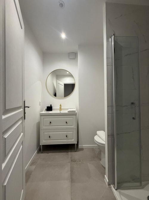 Salle de bain moderne de l'Appart Hôtel Panambi avec vasque blanche, miroir rond et douche à l'italienne à Chevilly-Larue