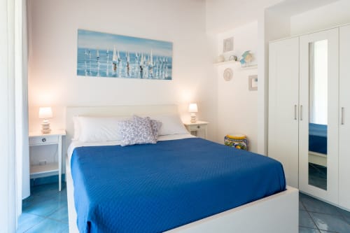 Camera da letto luminosa a CefalHome Roma con copriletto blu, mobili bianchi, opera d'arte con barca a vela e arredamento costiero