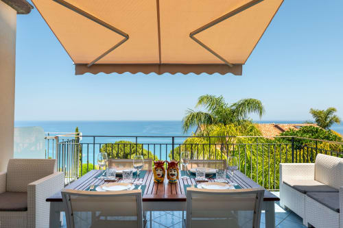CefalHome luxury terrace in Roma overlooking the sea with dining table, tropical plants, and beige umbrella