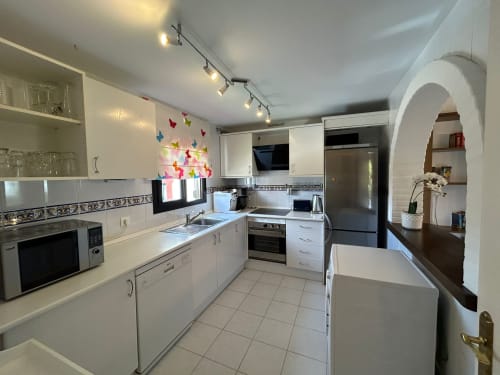 Cocina moderna blanca con electrodomésticos de acero inoxidable, arte decorativo de mariposas en pared y pasillo hacia sala de estar