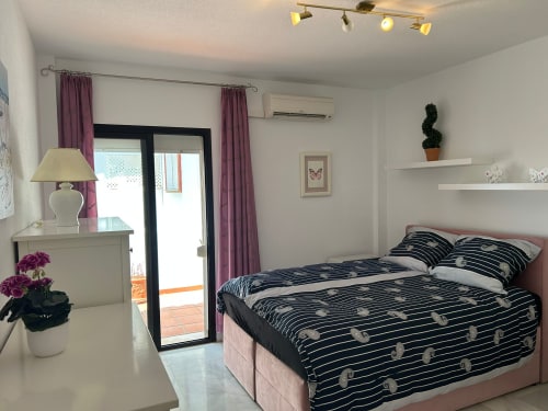 Dormitorio moderno en apartamento SACORO en Marbella con cama a rayas, balcón con vistas al mar, estantería blanca y cortinas moradas