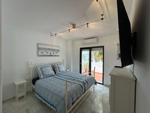 Dormitorio luminoso en apartamento SACORO, Marbella, con ropa de cama de rayas azules, iluminación moderna y acceso a terraza con vistas al mar