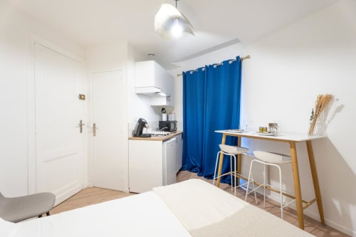 Modern studio apartment at Wellkhome Bordeaux with compact kitchen, blue curtains, work desk, and bright minimalist decor