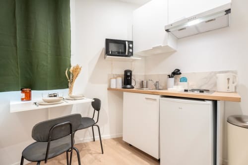 Modern smart studio kitchen at Wellkhome Bordeaux with white cabinets, wooden shelf, microwave, and dining area with green