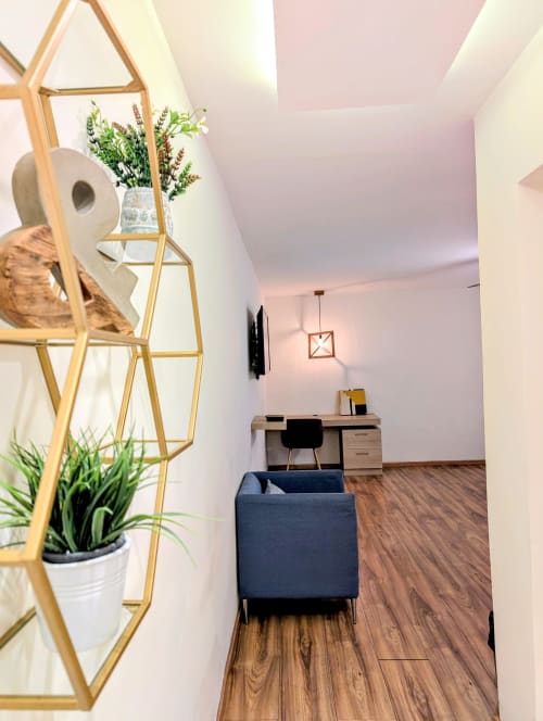Entrada moderna de estudio en Casa Donceles con estanterías doradas, plantas, escritorio, silla azul marino y piso de madera en México.