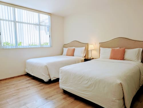 Dormitorio luminoso con dos camas queen, cabeceros tapizados en beige, almohadas coral, piso de madera y amplia ventana en Casa
