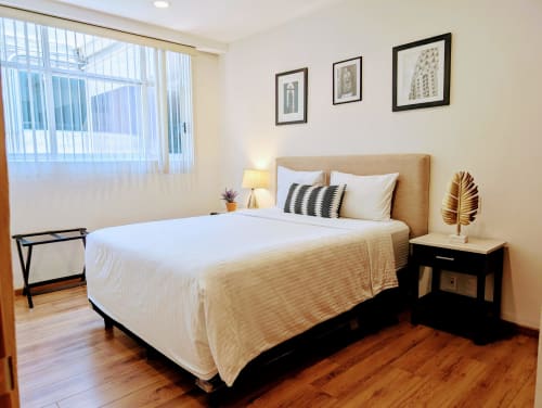 Dormitorio luminoso en Casa Donceles, Ciudad de México, con cama queen, mesita de noche de madera, ventanales grandes y arquitectura enmarcada