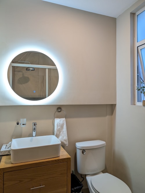 Baño moderno del penthouse Casa Donceles con espejo redondo iluminado, lavamanos blanco, inodoro y tocador de madera