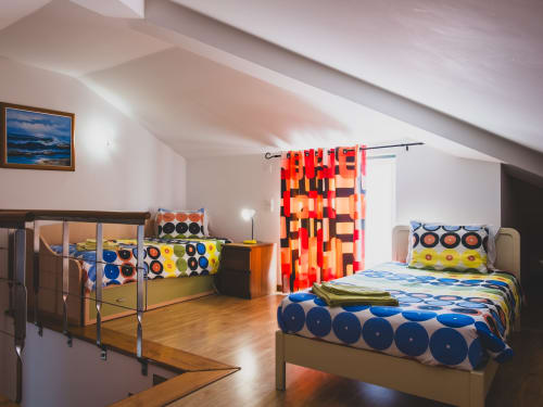 Quarto luminoso no sótão com duas camas, roupa de cama colorida estampada, assoalho de madeira e cortinas vermelhas na villa de Bombarral.