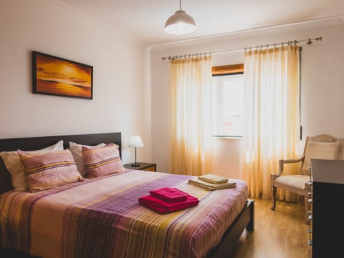Quarto luminoso em moradia portuguesa com roupa de cama às riscas, cortinados com vista para o mar e comodidades modernas na Home Alliance Bombarral