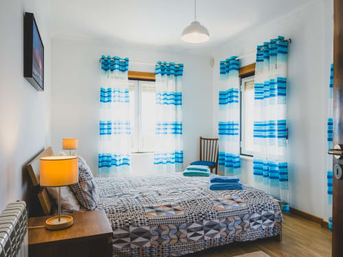 Quarto luminoso com cortinas listradas em azul e branco, roupa de cama estampada, luminária laranja e piso de madeira em Fabulous Beach