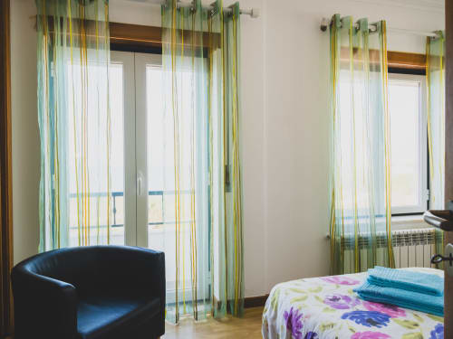 Quarto em villa atlântica com vista ao mar, cortinas verdes listradas, poltrona azul, roupa de cama floral e móveis modernos.
