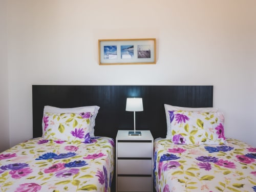 Camas gêmeas com roupa de cama floral colorida no quarto de villa à beira-mar, cabeceira preta, criado branco com abajur, decoração costeira emoldurada