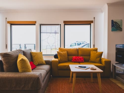 Sala de estar de villa moderna com vista para o mar, sofá mostarda, poltrona cinzenta, mesa de café em madeira e decoração em tons neutros aconchegantes