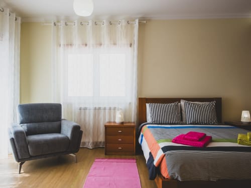 Quarto luminoso na villa Baleal Sunset Residence com poltrona cinza, cama de madeira com almofadas listradas, mantas coloridas e