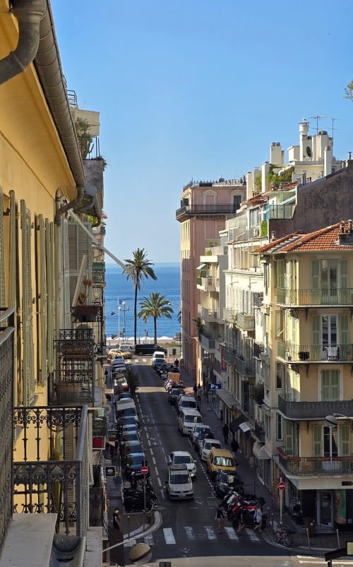 Street view from Hôtel De La Fontaine in Nice showing Mediterranean sea, palm trees, and colorful buildings