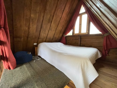 Cozy A-frame chalet bedroom with white bedding, wooden beams, triangular window, red curtains, and mountain views at Chalets