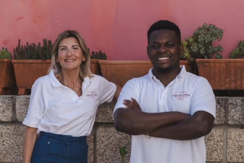 Dos amables empleados de Casa das Jardas posan frente a una pared rosa con macetas de barro.
