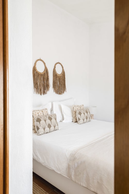 Quarto minimalista na Casa das Jardas com roupa de cama branca, almofadas estampadas e tapeçarias tecidas com franjas