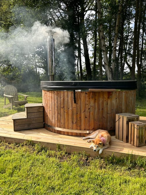 Outdoor wooden hot tub with smoking chimney, dog resting on deck, Adirondack chair, forest setting at Domaine de Blanvatel,